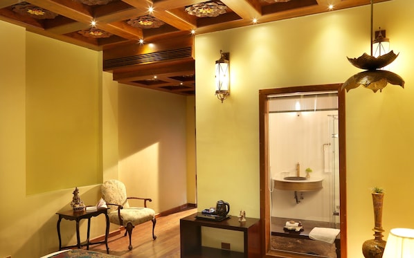 Una habitación con techo de madera, una silla, una mesita con una tetera, un espejo y un baño visible a través de una puerta de cristal.