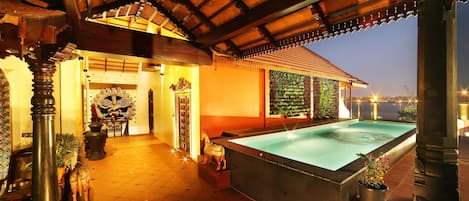 Zona de piscina con suelo de baldosas, una piscina grande y una sección cubierta con techo de madera.