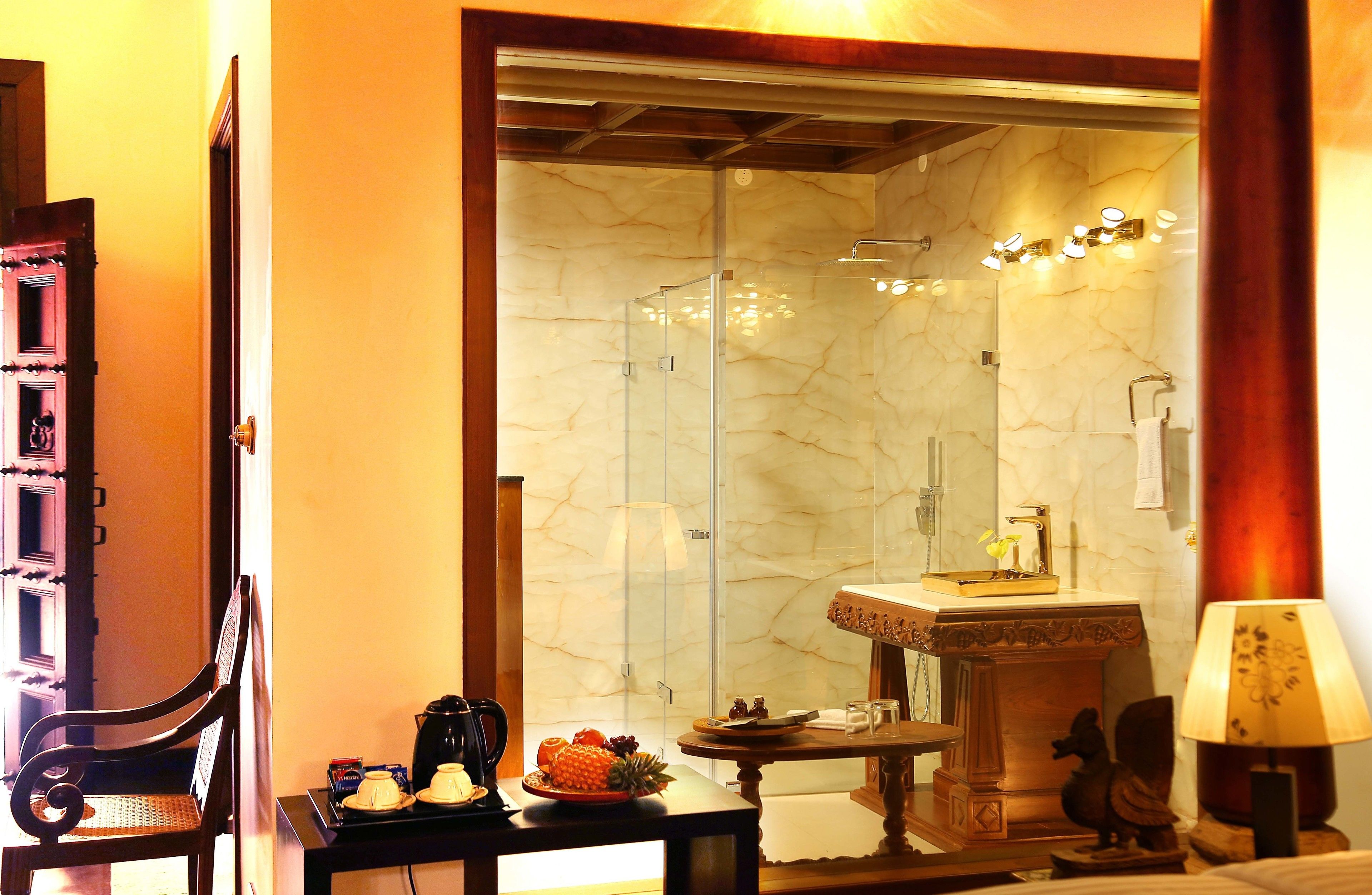 Une salle de bain d’hôtel avec un mur en marbre, une douche en verre, un lavabo avec un receveur décoratif et une petite table avec des articles de toilette.