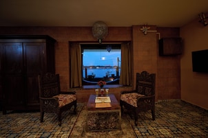 A room with a view of a bridge and water, two ornate chairs, a table with a vase and a plate, and a clock on the wall.