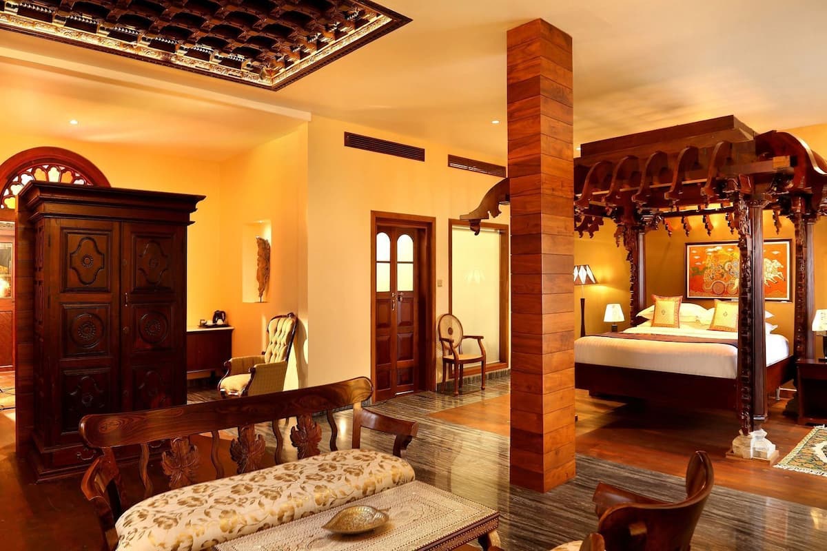 A hotel room with a bed, a wooden canopy bed, a sitting area with a bench and chairs, a large wooden wardrobe, and a decorative wall piece.