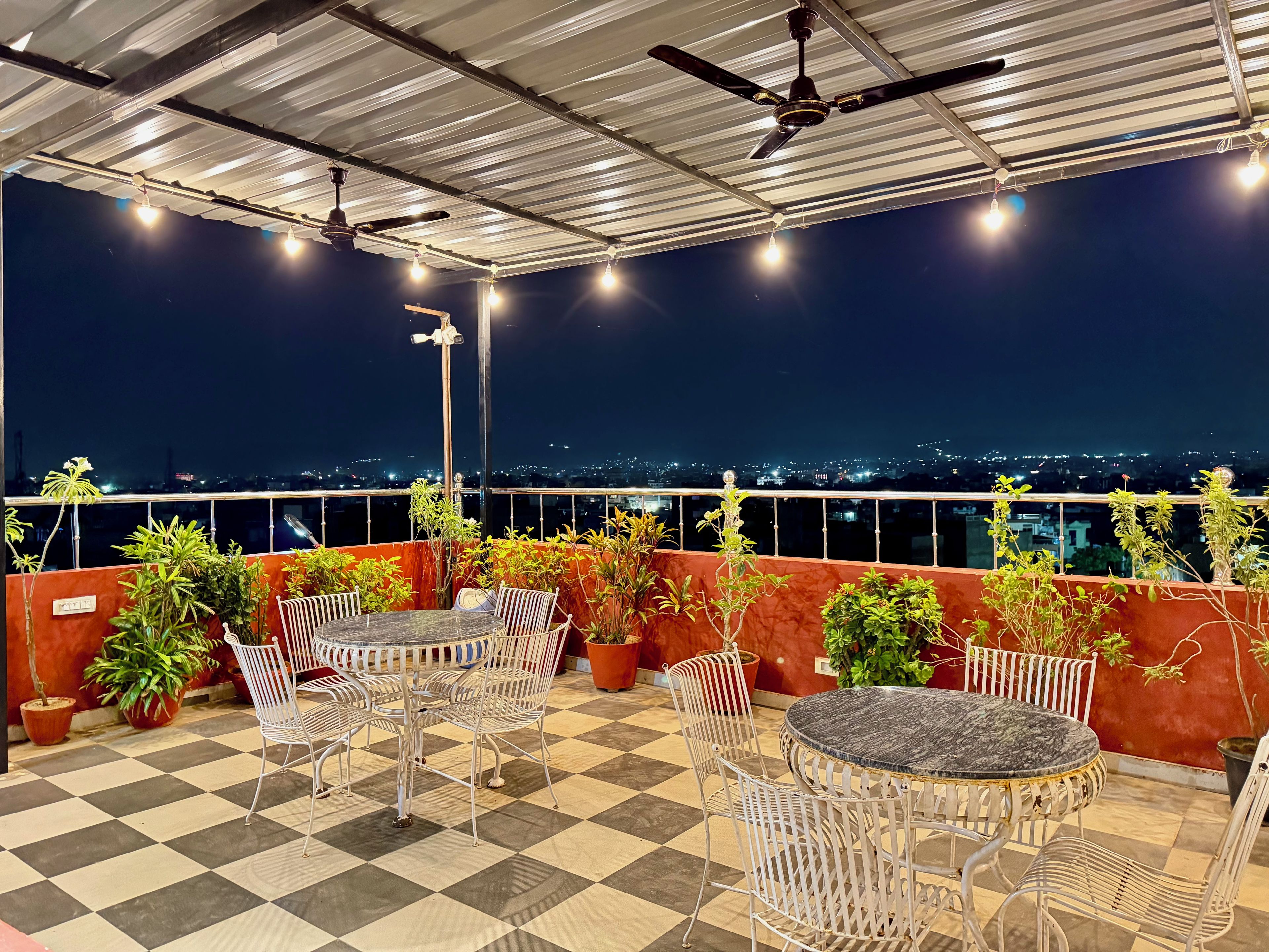 A rooftop terrace with a checkered floor, metal chairs, a round table, potted plants, and a cityscape view at night.