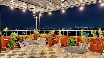 Una terraza en la azotea con piso a cuadros, sillas de metal, una mesa redonda, plantas en macetas y vista panorámica de la ciudad de noche.