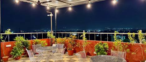 Una terraza en la azotea con piso a cuadros, sillas de metal, una mesa redonda, plantas en macetas y vista panorámica de la ciudad de noche.