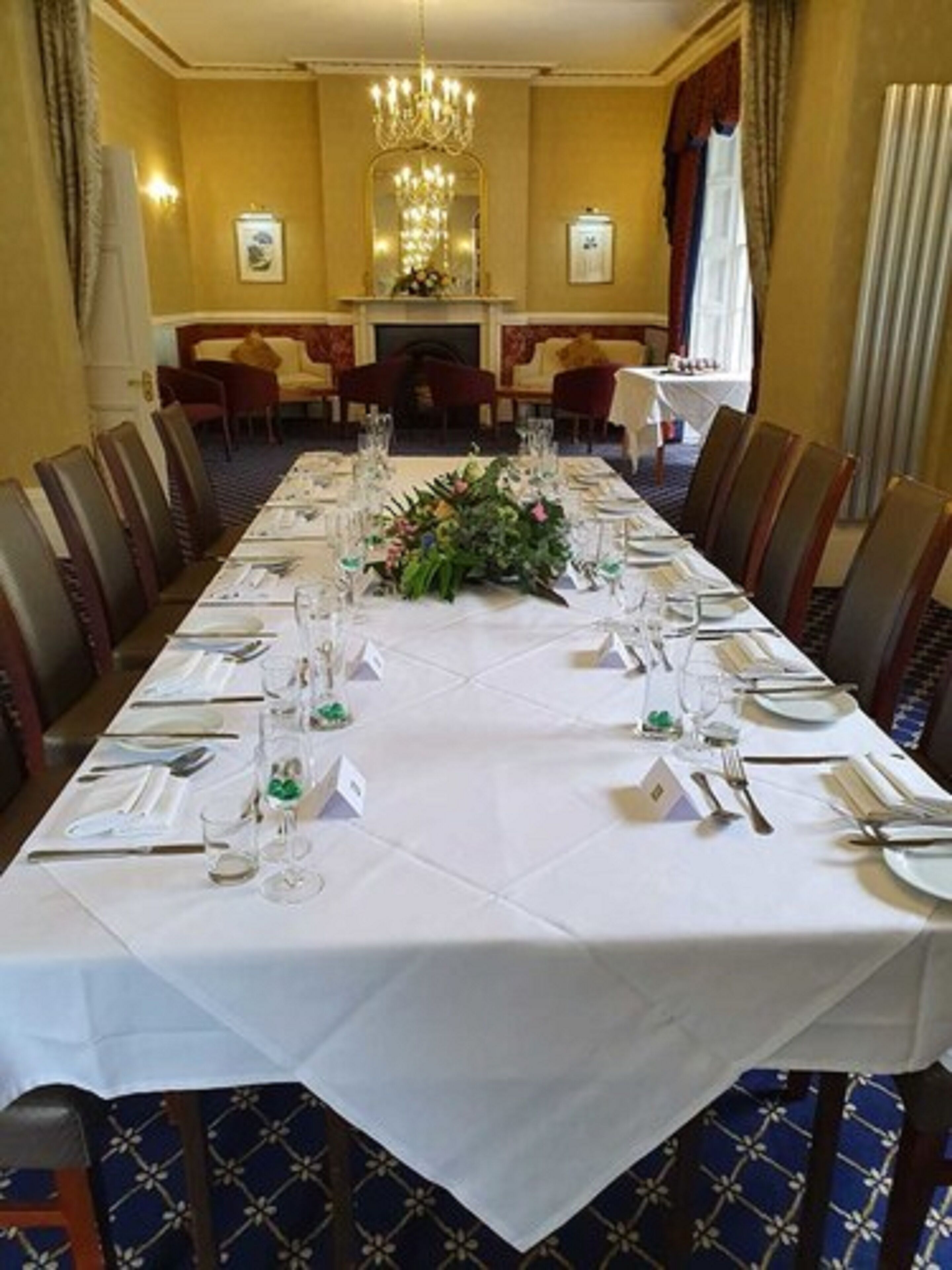 Een lange eettafel gedekt voor een formeel diner, met aan weerszijden stoelen, een kroonluchter erboven en een open haard op de achtergrond.