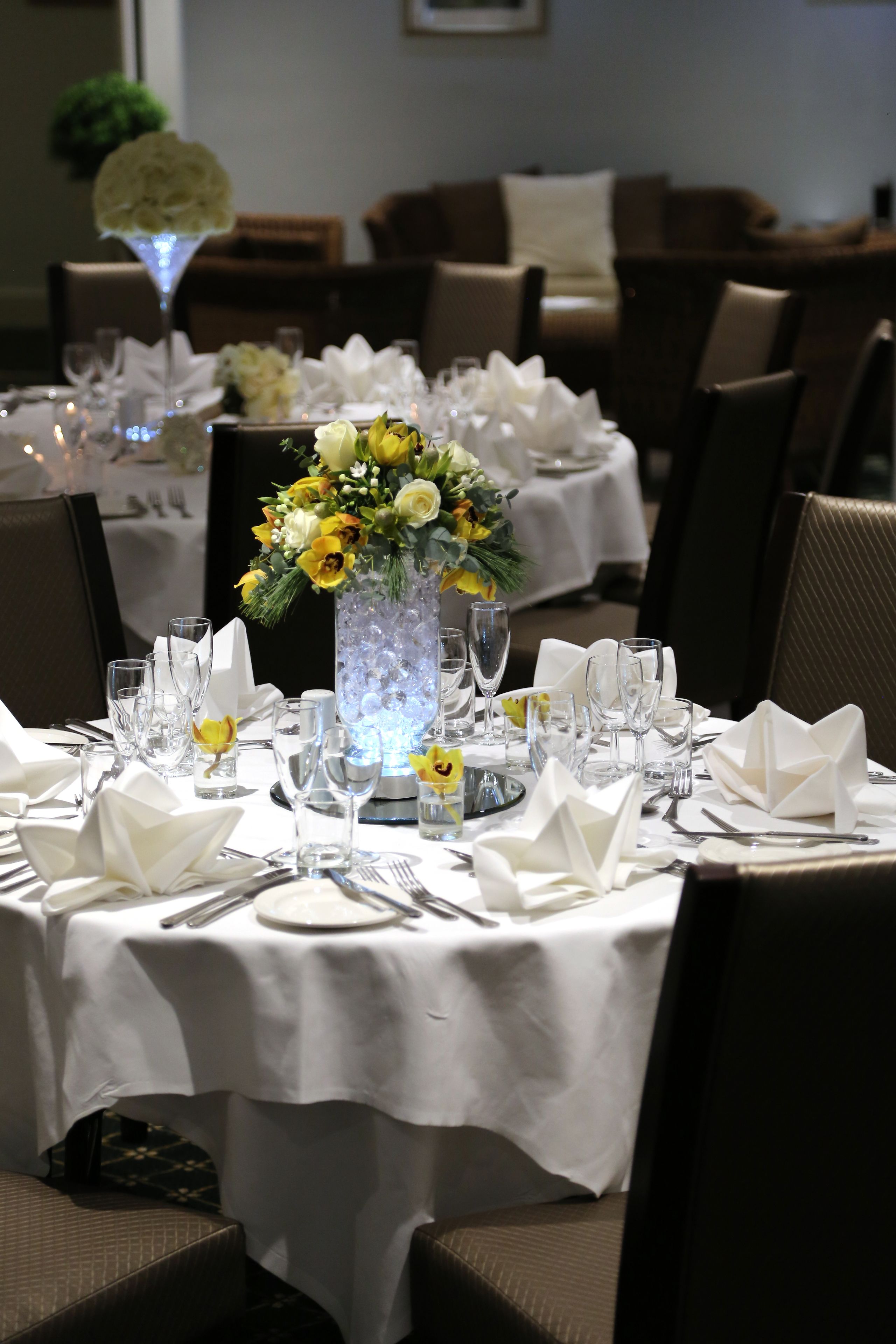 A dining table set for a formal meal with white tablecloths, folded napkins, and a floral centerpiece.