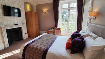Ein Hotelzimmer mit Bett, Wandfernseher, Kamin, Fenster mit Vorhängen und einem kleinen Tisch mit Stuhl.