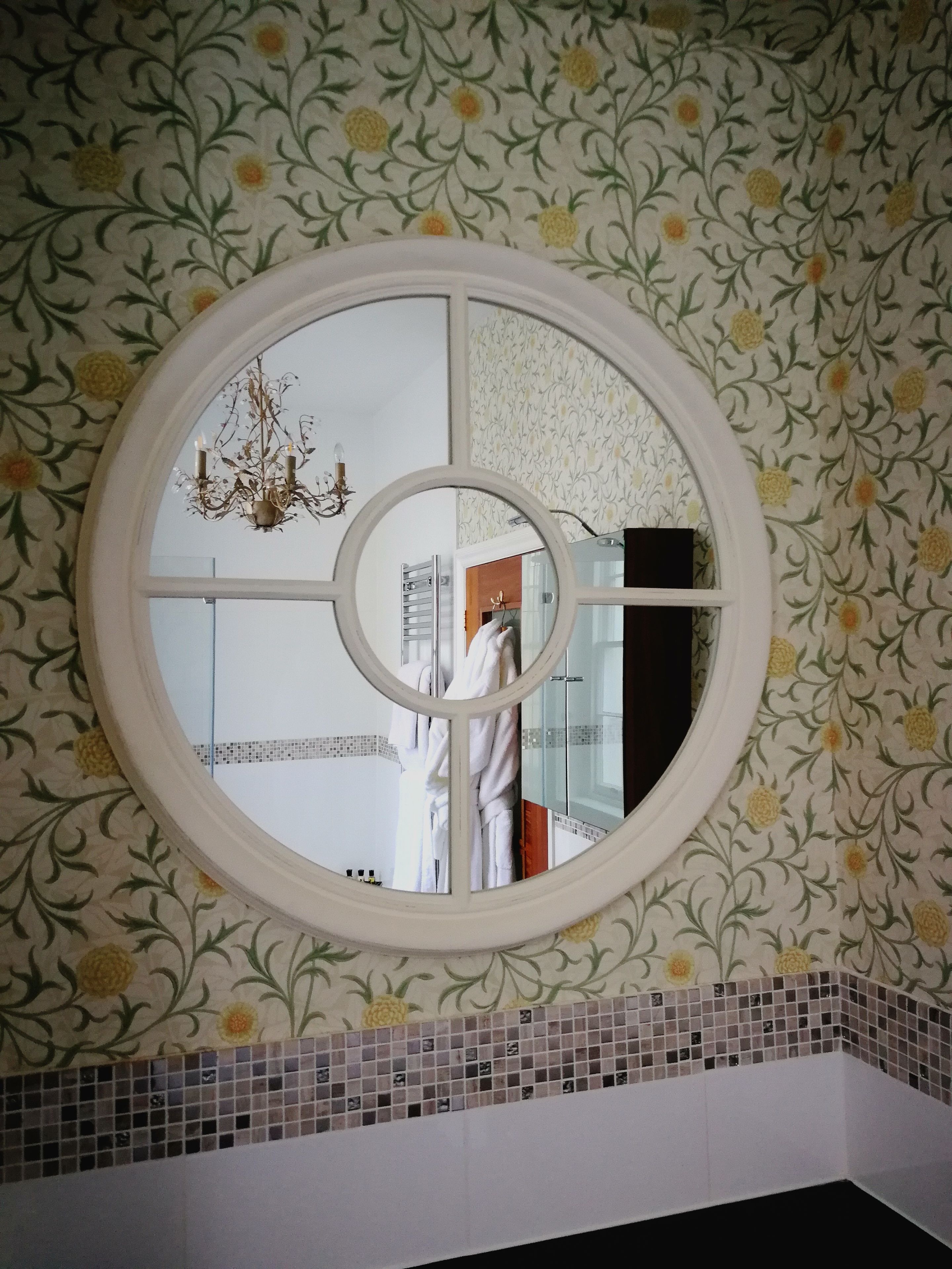 Un baño con papel tapiz floral, un espejo redondo y una araña.
