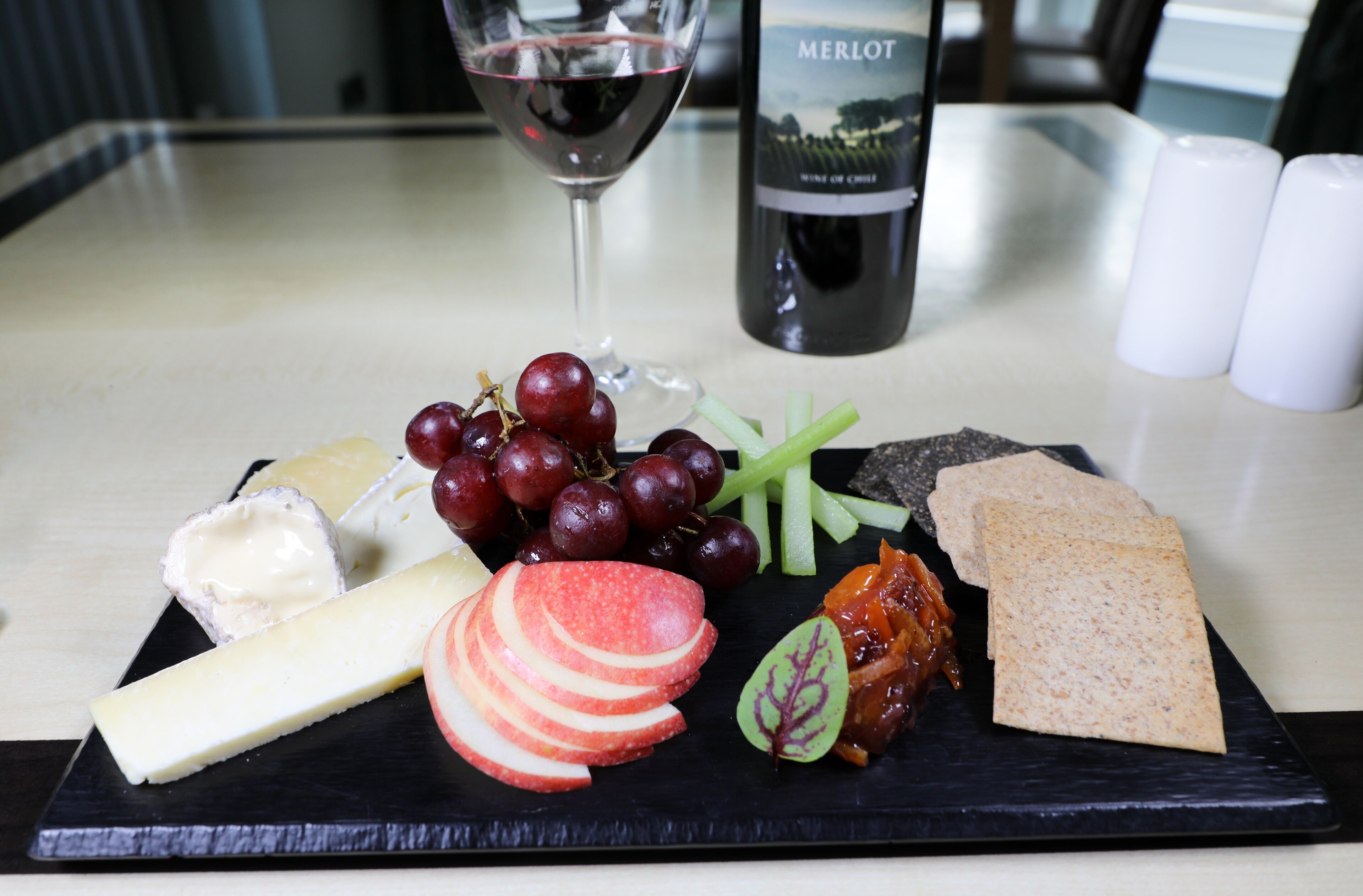 A black slate board with cheese, grapes, apple slices, and crackers on a table. A bottle of Merlot wine and a glass of red wine are also visible.