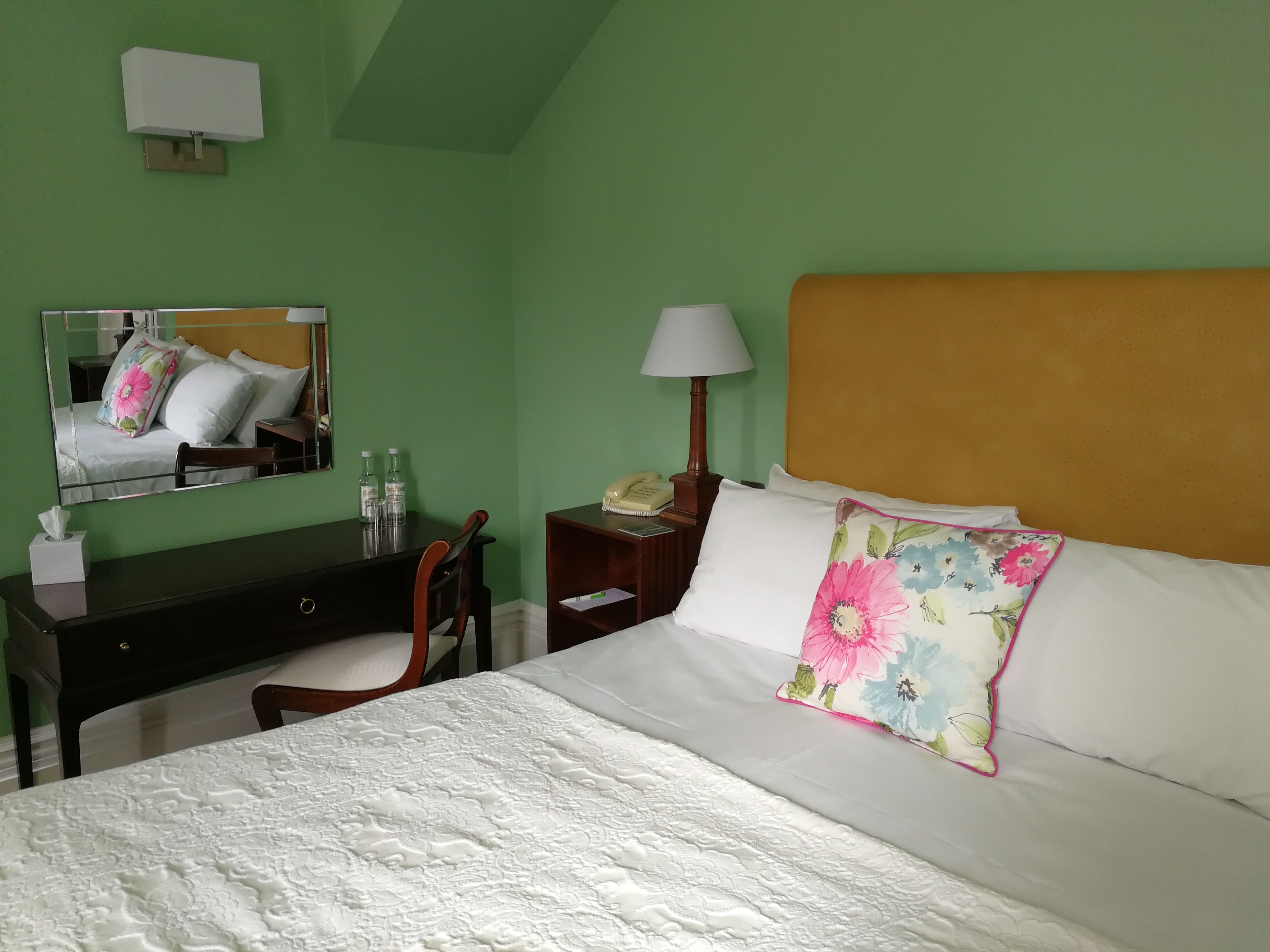 A hotel room with a bed, a desk, a chair, a nightstand, a lamp, and a mirror.