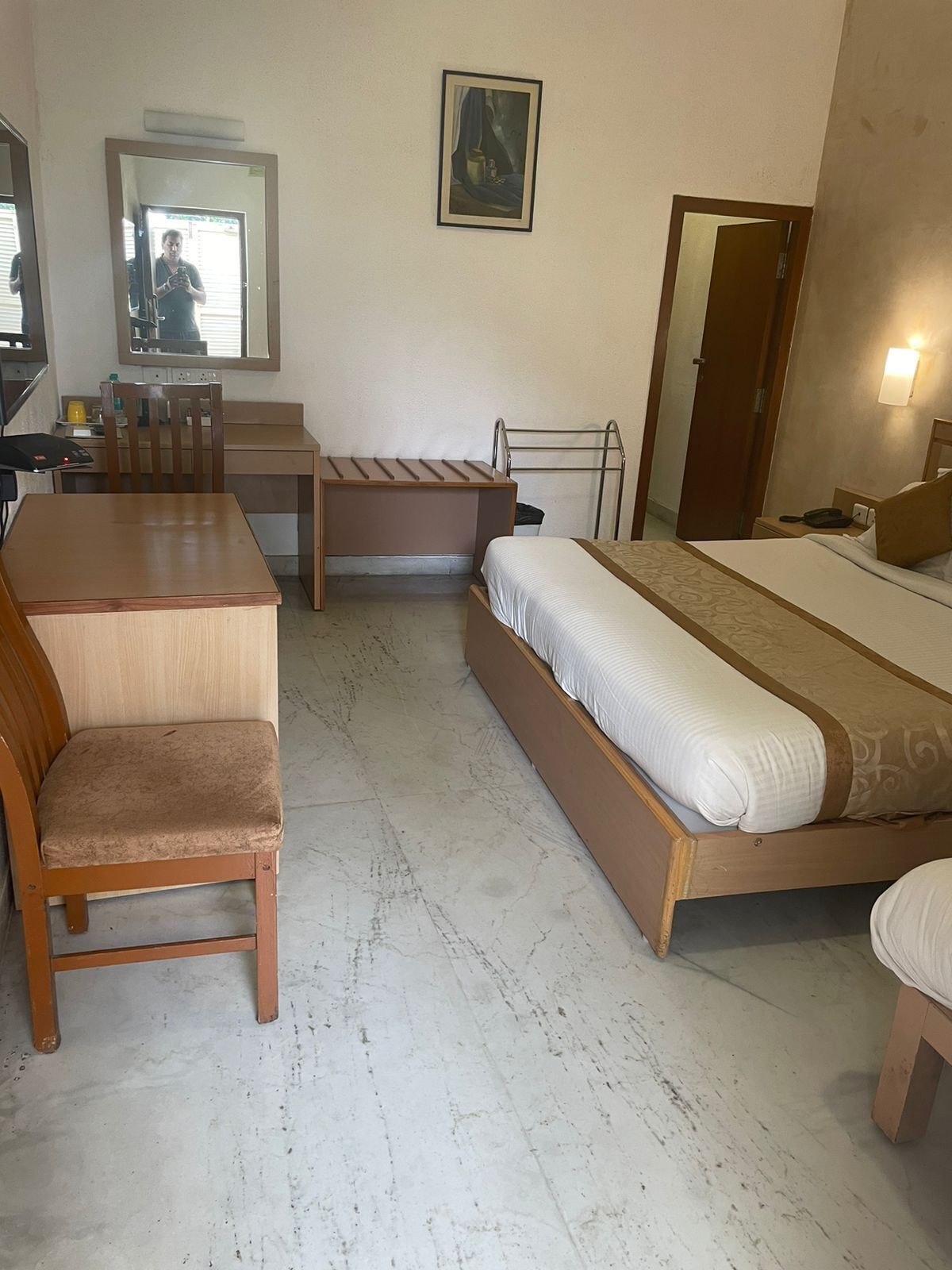 A compact hotel room with a bed, a desk, a chair, a small table, a mirror, and a bedside lamp.