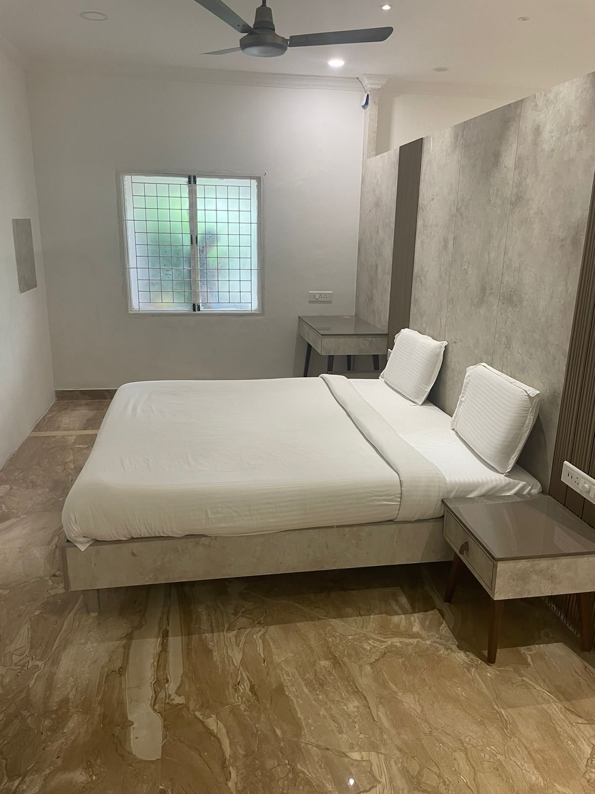 A modern bedroom with a large bed, a nightstand, a desk, and a window with blinds.