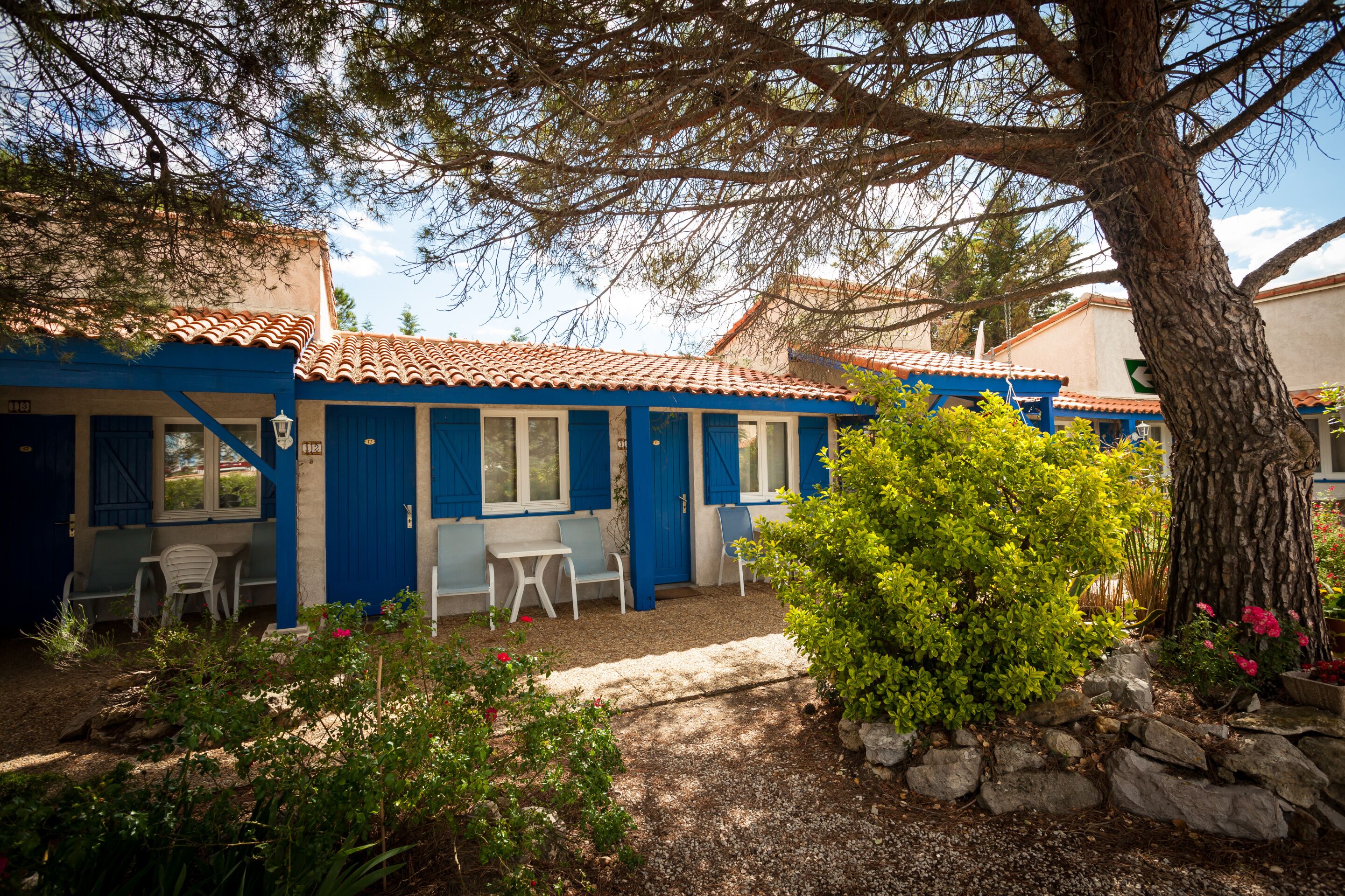 Un edificio con puertas y ventanas azules, un patio con sillas blancas y un jardín con rocas y plantas.