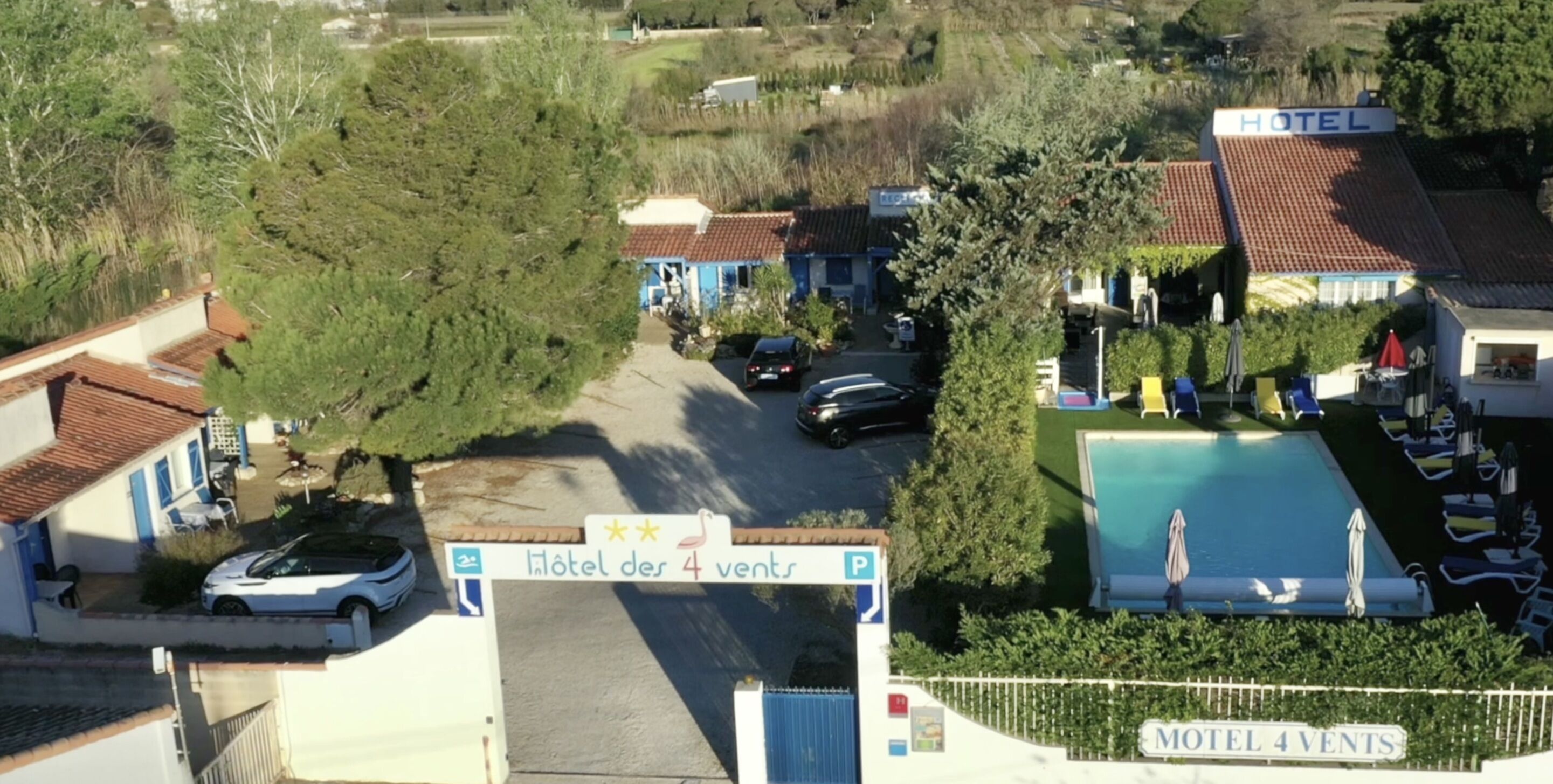A hotel with a swimming pool, a parking area, and a sign indicating 'Hotel des 4 Vents'.