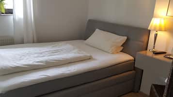 Una cama bien hecha con sábanas blancas, un cabecero gris y una mesita de noche con una lámpara.