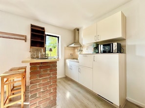 A kitchen with a brick wall, white cabinets, a wooden bar stool, and a window with a view of greenery. - Landhotel Linden am Venekotensee (Niederkruechten)