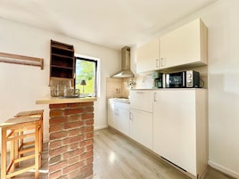 Una cocina con pared de ladrillo, armarios blancos, un taburete de madera y una ventana con vistas a follaje.