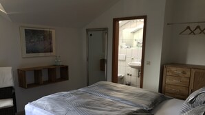 Un dormitorio con cama, cómoda de madera, mesita de noche con vasos, un cuadro en la pared y un baño visible a través de una puerta abierta.