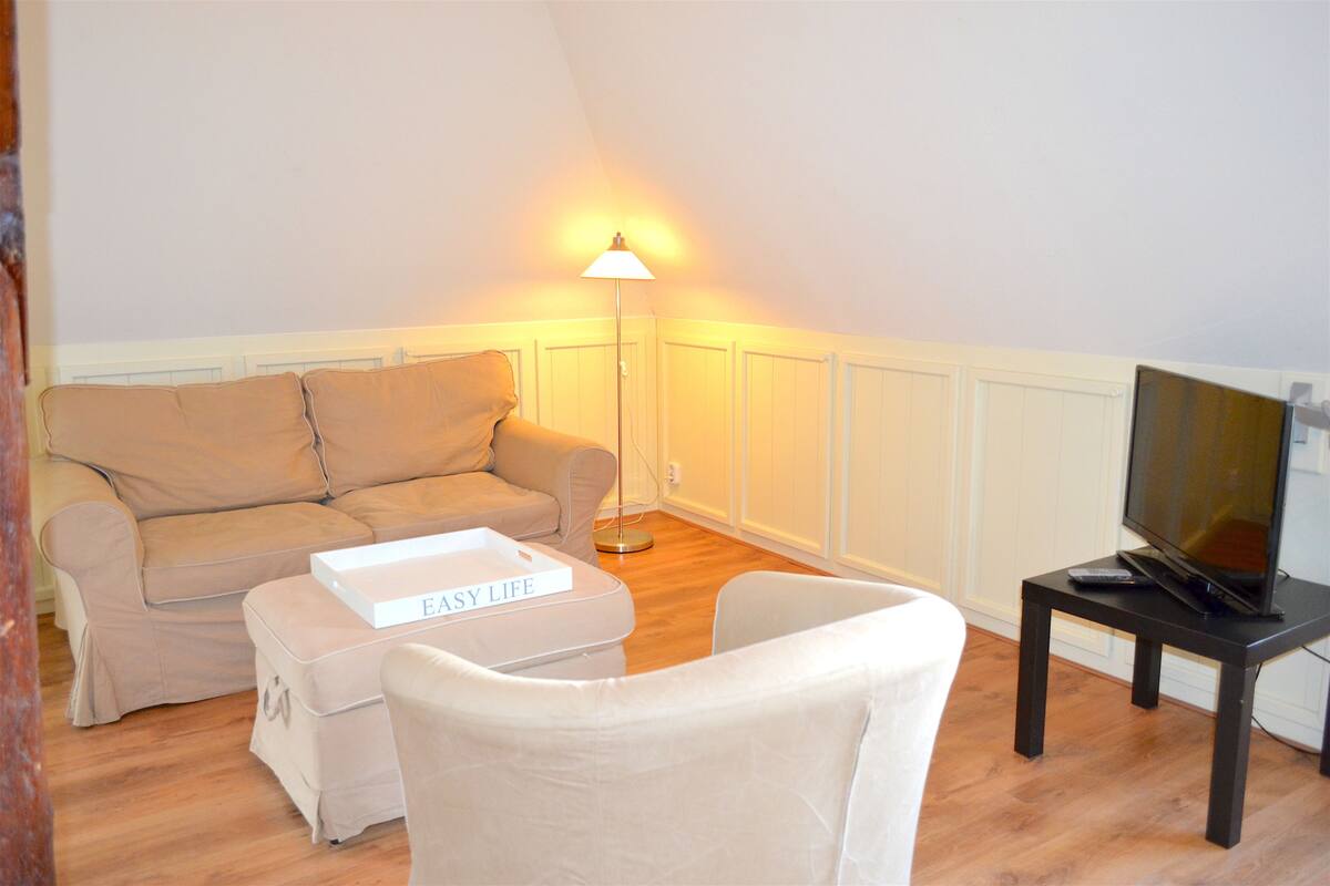 A living room with a beige sofa, a white armchair, a small table with a remote control, and a TV on a stand.