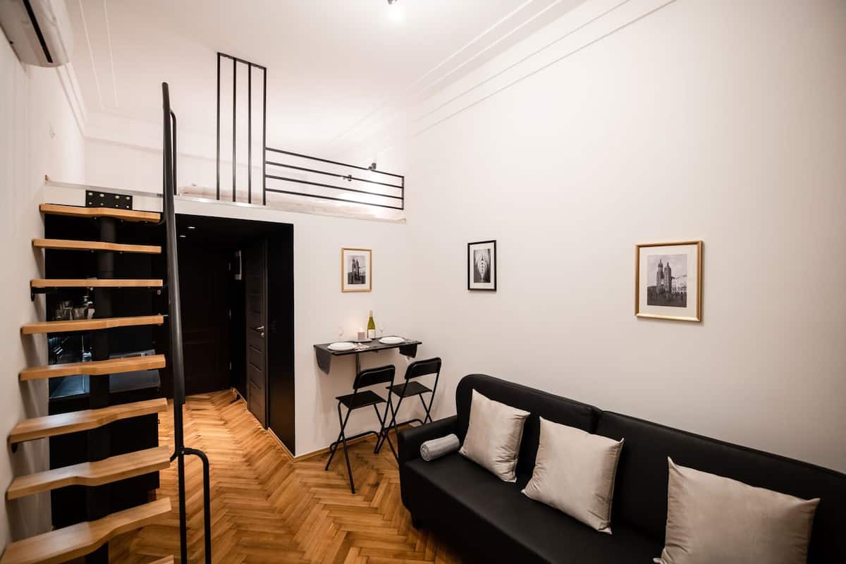 A compact living space with a black sofa, a dining table with chairs, a wooden staircase, and framed pictures on the walls.