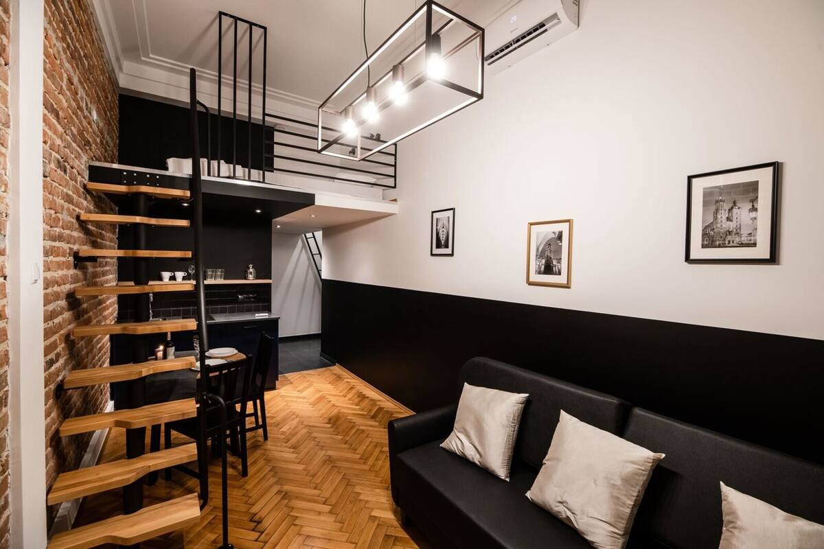 A modern loft-style room with a herringbone wood floor, a black sofa, a wooden ladder, and a mezzanine with a staircase.