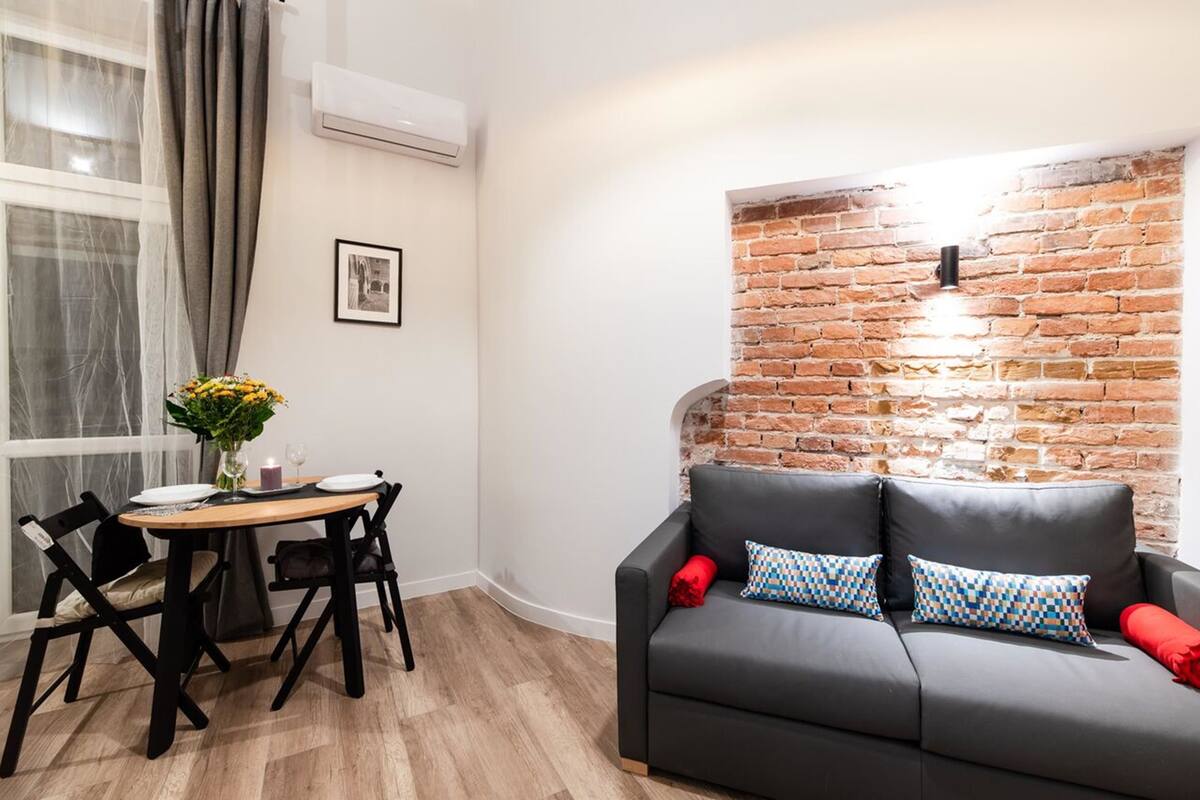 A cozy living area with a brick accent wall, a grey sofa with patterned cushions, and a dining table set for two.