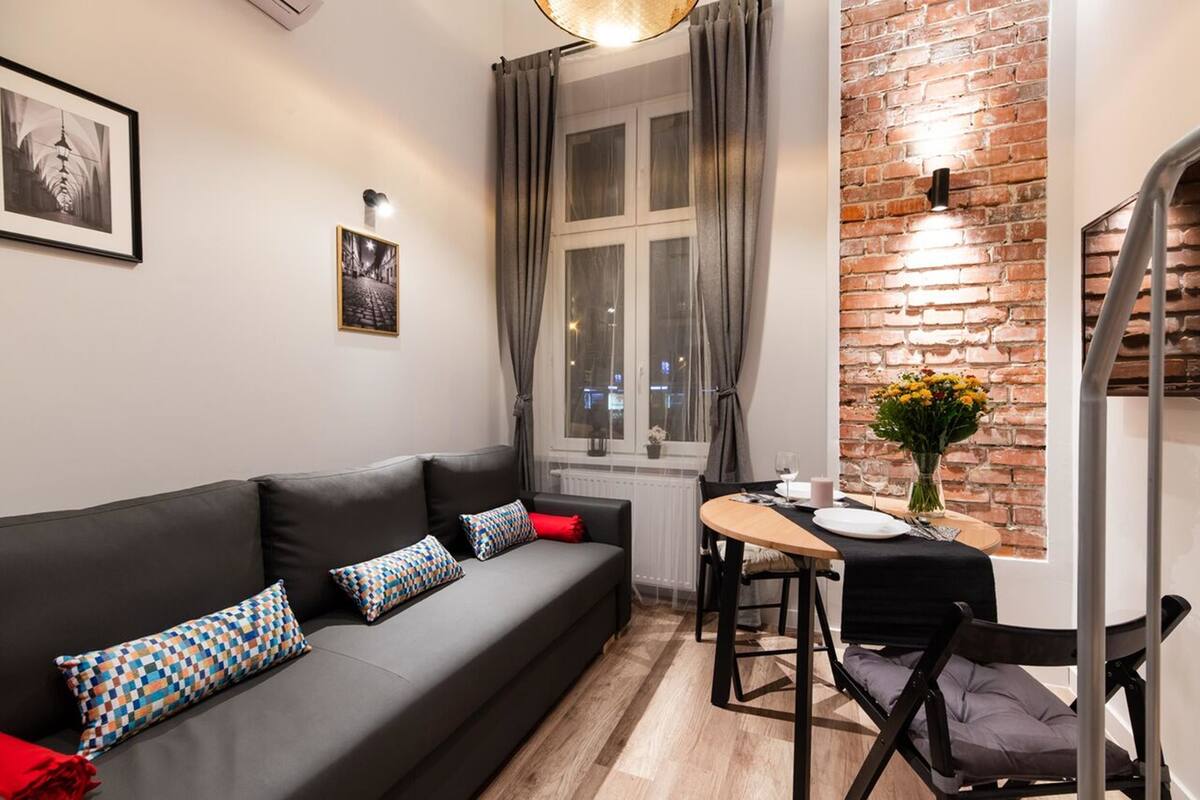 A modern living room with a black sofa, patterned cushions, a dining table with chairs, a vase of flowers, and a brick wall.