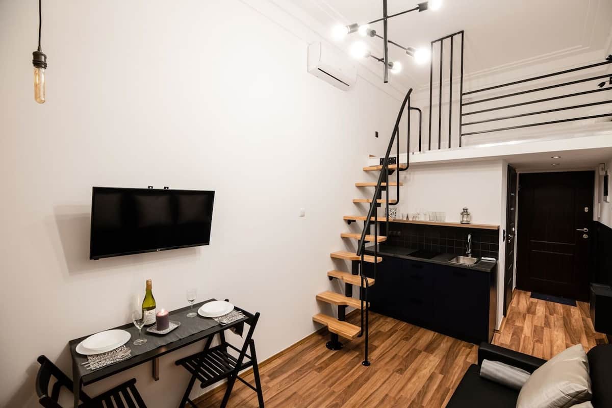 A compact living space with a dining area, a TV mounted on the wall, a staircase leading to an upper level, and a kitchen area.