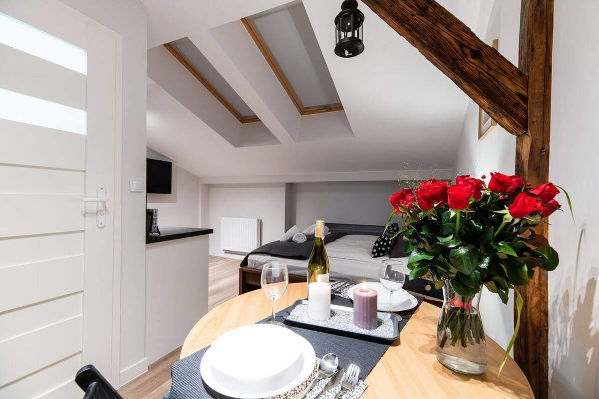 A modern bedroom with a skylight, a bed with a black and white polka dot pillow, a bedside table with a wine bottle, candles, and a vase of red roses.