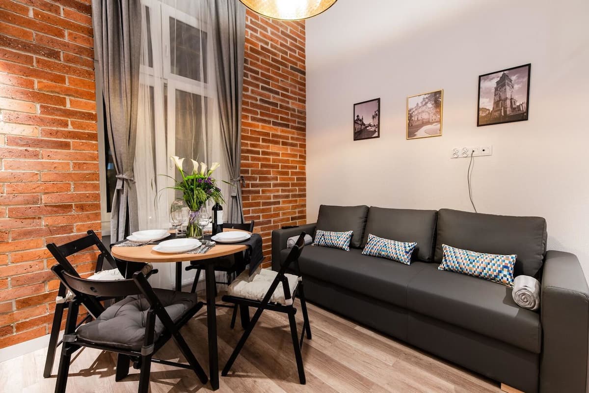 A modern living room with a brick wall, a black sofa with patterned cushions, a dining table with chairs, and a window with curtains.