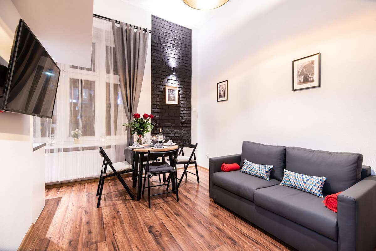 A modern living room with a black leather sofa, a dining table with chairs, a TV mounted on the wall, and a large window with curtains.