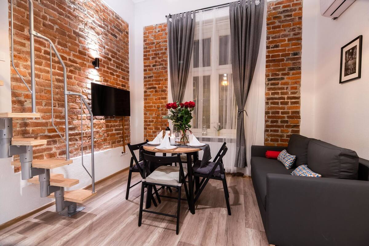 A cozy living room with a brick wall, a dining table with chairs, a black sofa, and a TV mounted on the wall.