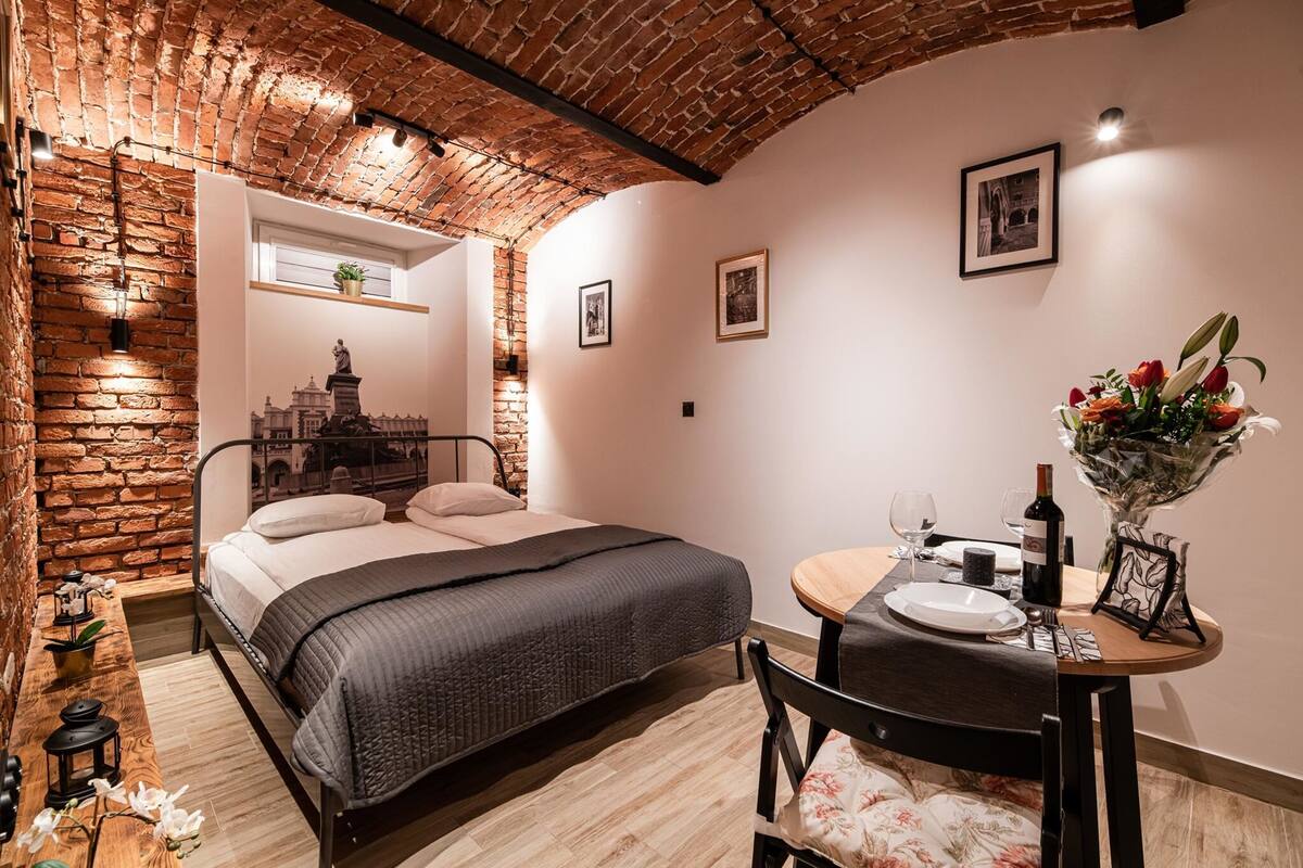 A cozy bedroom with a brick wall, a bed, a small table with a vase of flowers, and a framed picture on the wall.