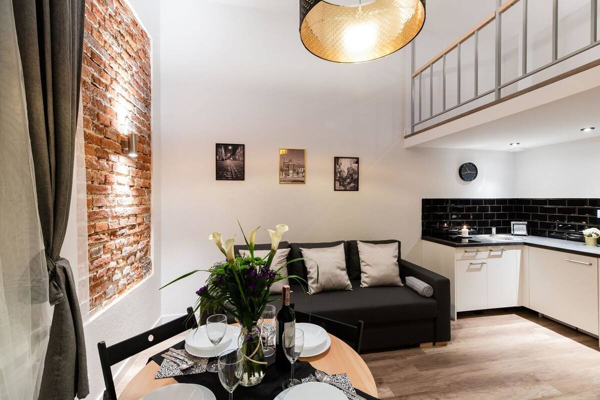 A modern dining area with a brick accent wall, a black sofa, and a kitchen in the background.