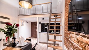 Un comedor moderno con una mesa de madera, sillas y un jarrón de rosas rojas. Una escalera conduce a un nivel superior con un muro de ladrillo y un televisor.