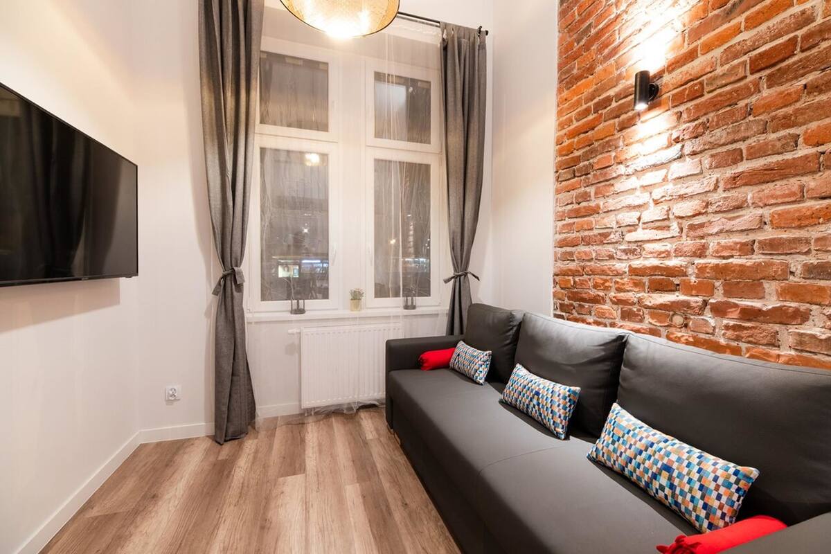 A modern living room with a black leather sofa, patterned cushions, a flat-screen TV, and a brick wall.