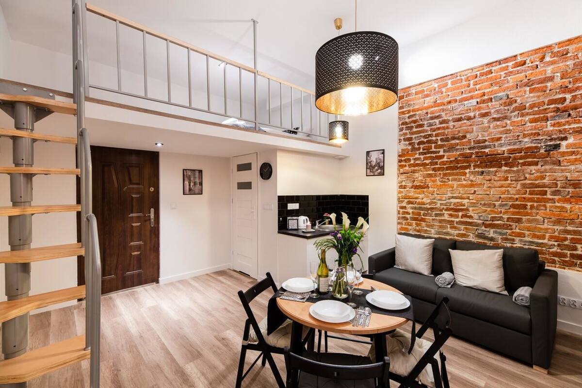 A modern living room with a dining area, a staircase, a brick wall, and a black leather sofa.
