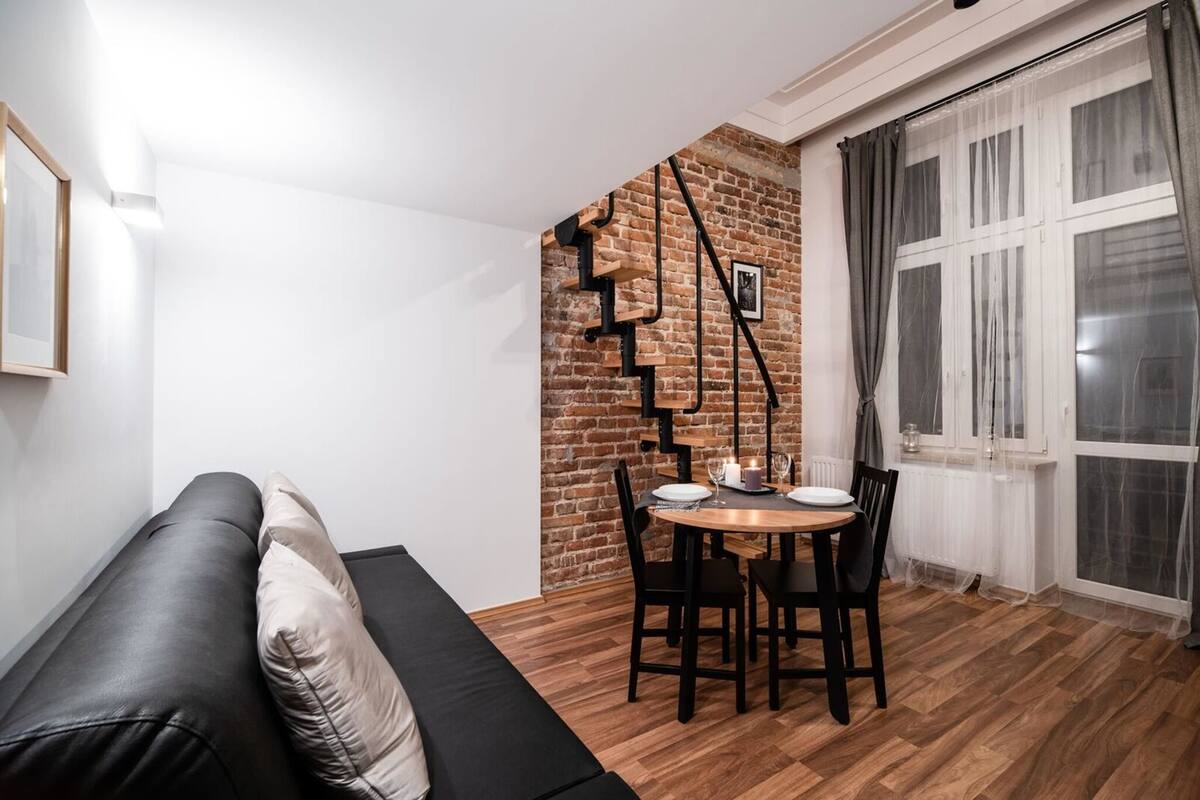 A cozy living area with a black sofa, a wooden dining table with chairs, a brick wall, and a window with curtains.