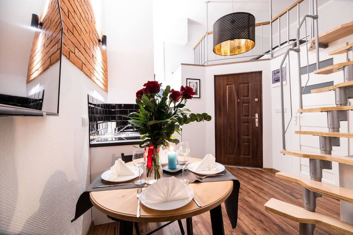 A dining area with a round table set for four, a vase with red flowers, and a modern staircase with wooden steps and metal railings.
