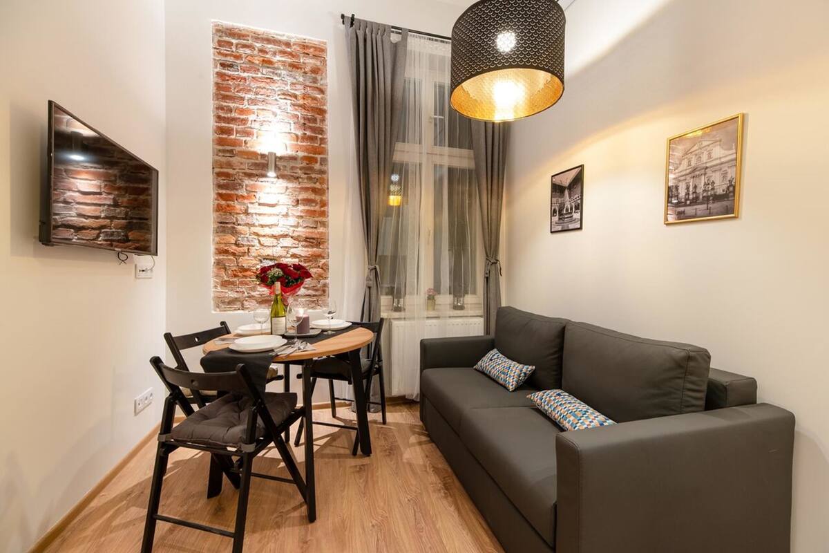 A compact living space with a dining area, a sofa, a brick accent wall, a large window with curtains, and a hanging light fixture.