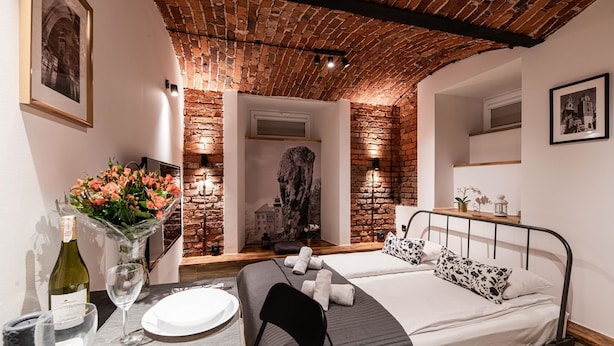 Habitación de hotel con pared de ladrillo, cama con estructura metálica negra, una mesita con un jarrón de flores y un cuadro en la pared.