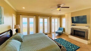 A bedroom with a bed, a chair, a TV, a fireplace, and large windows with curtains. - Portofino - Private Beachfront House 3 Bedroom Home by RedAwning (South Padre Island)