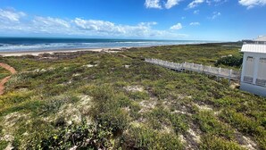 A coastal landscape with a beach, a white building with a balcony, and a white fence leading to the beach. - Portofino - Private Beachfront House 3 Bedroom Home by RedAwning (South Padre Island)
