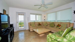 A living room with a sectional sofa, a coffee table, a TV on a stand, and a ceiling fan. - Seville Unit #201 3 Bedroom Condo by RedAwning (South Padre Island)