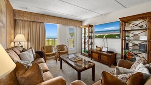 A spacious living room with a large window offering a view of the beach, a comfortable sofa set, a wooden coffee table, and a decorative shelf with a beach-themed sign.