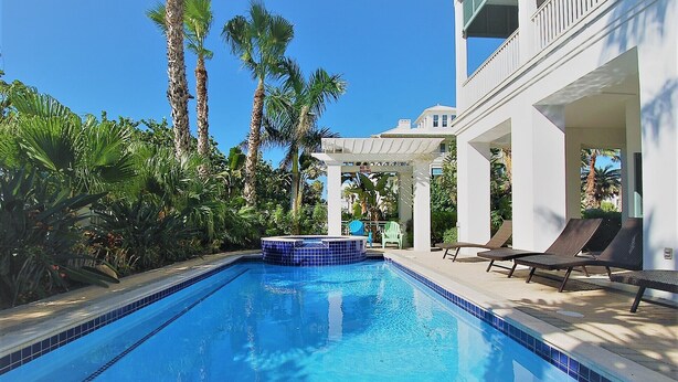 Piscina com espreguiçadeiras e hidromassagem, cercada por palmeiras e um edifício branco.