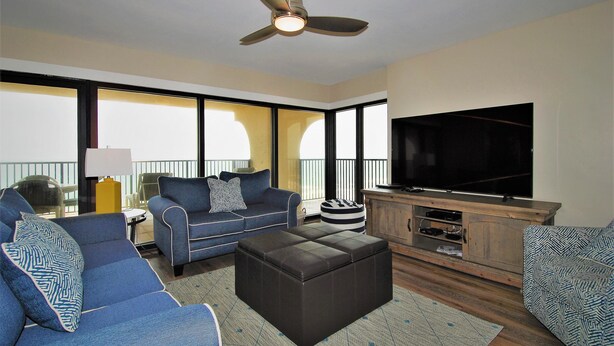 A modern living room with a large TV, comfortable seating, and a ceiling fan. - Ocean Vista Unit #1201 3 Bedroom Condo by RedAwning (South Padre Island)