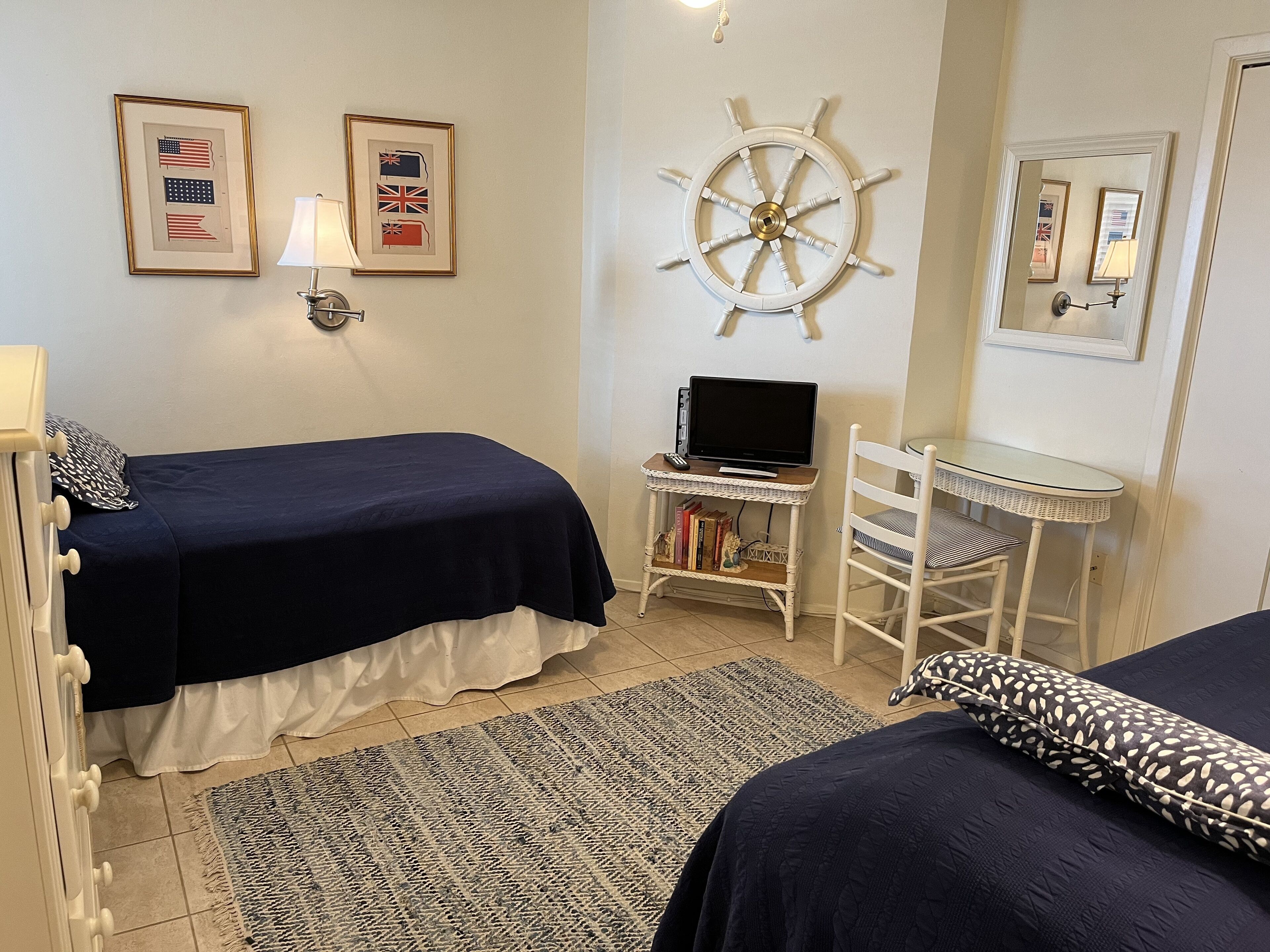 A bedroom with two beds, a desk with a computer, a ship's wheel decoration, and framed flags on the wall.