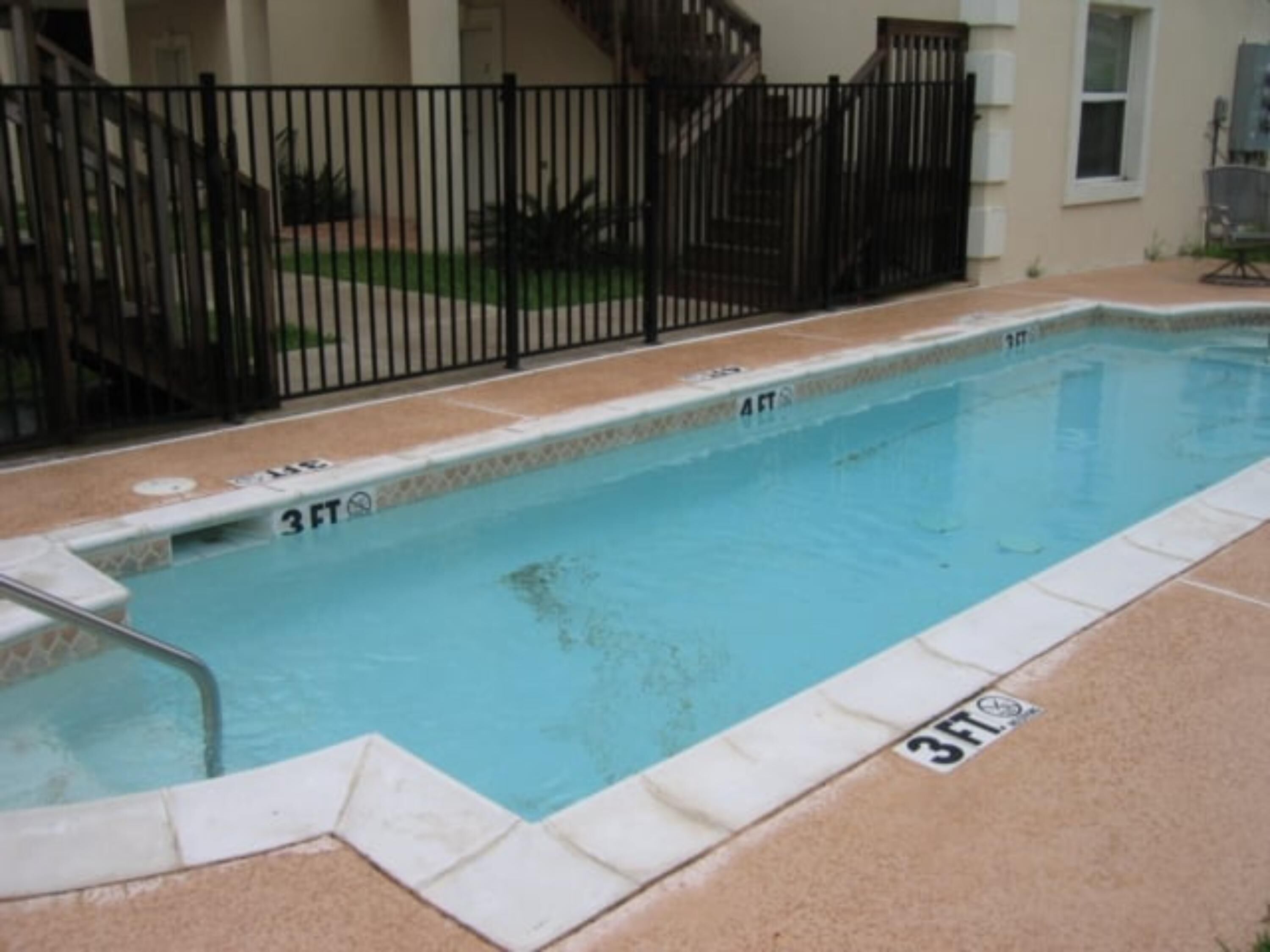 Ein rechteckiger Swimmingpool mit einem schwarzen Metallzaun, gefliesten Rändern und Tiefenmarkierungen für 3 und 4 Fuß.