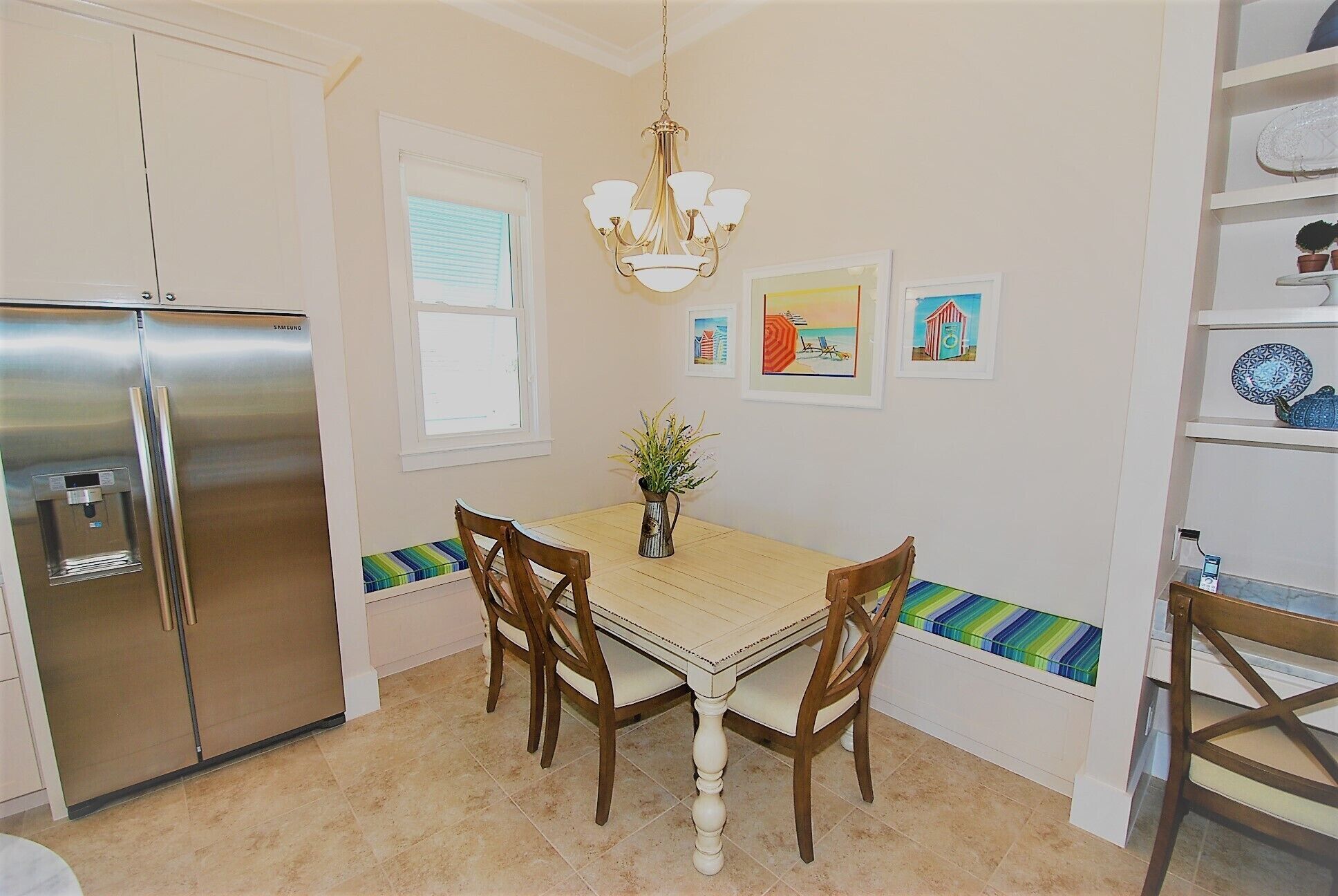 A kitchen with a stainless steel refrigerator, a dining table with four chairs, a chandelier, and a window with blinds.